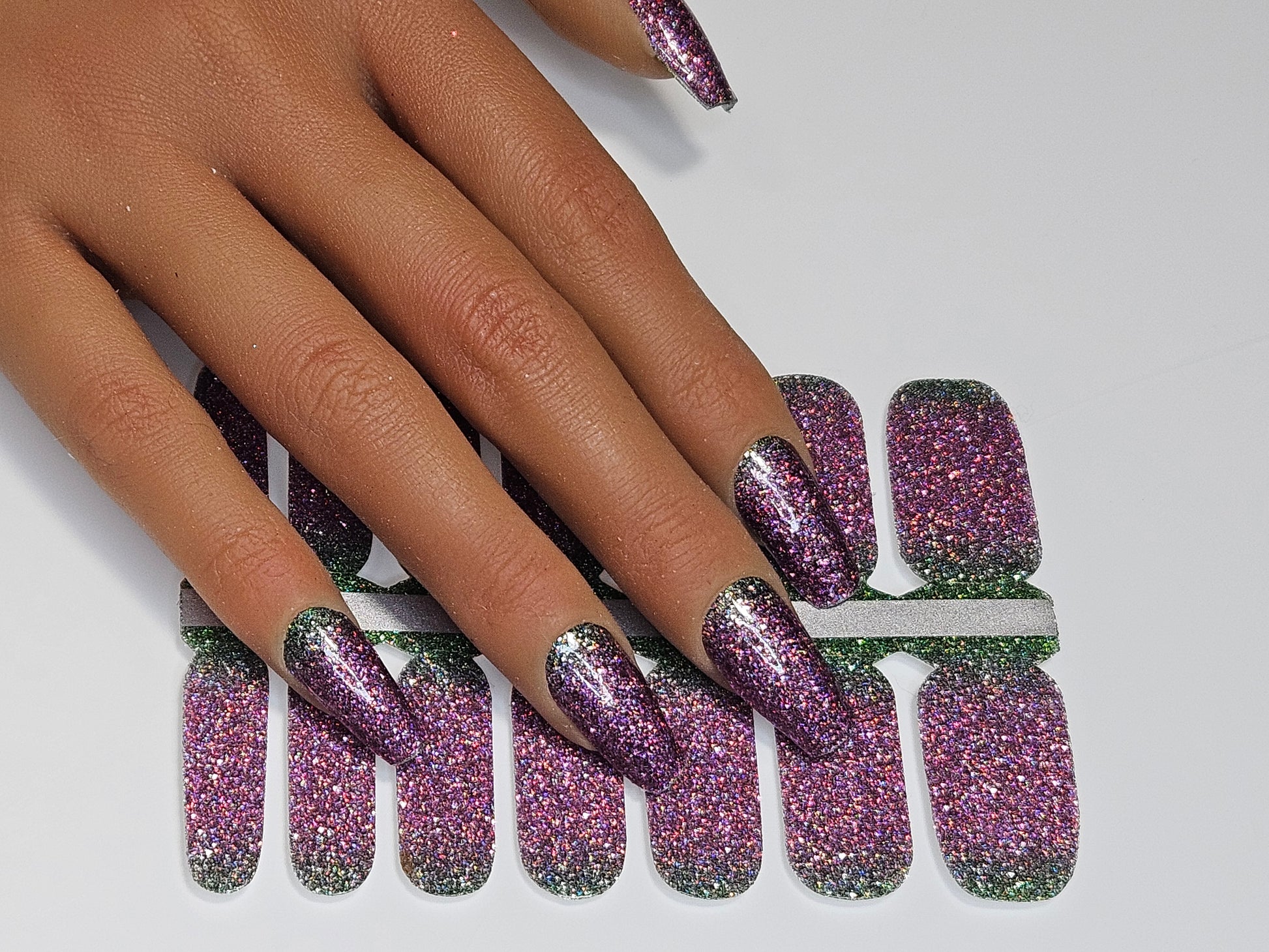Hand with purple and green glittery nail wraps on against a white background